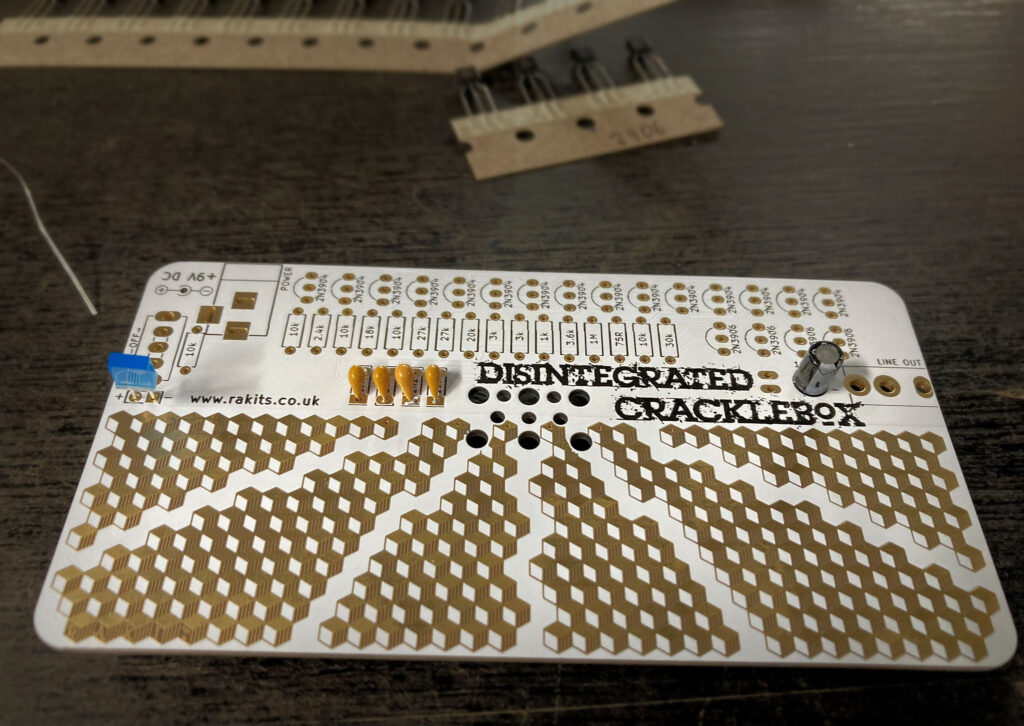 A white PCB labelled Disintegrated Cracklebox with capacitors and an LED now soldered in place.